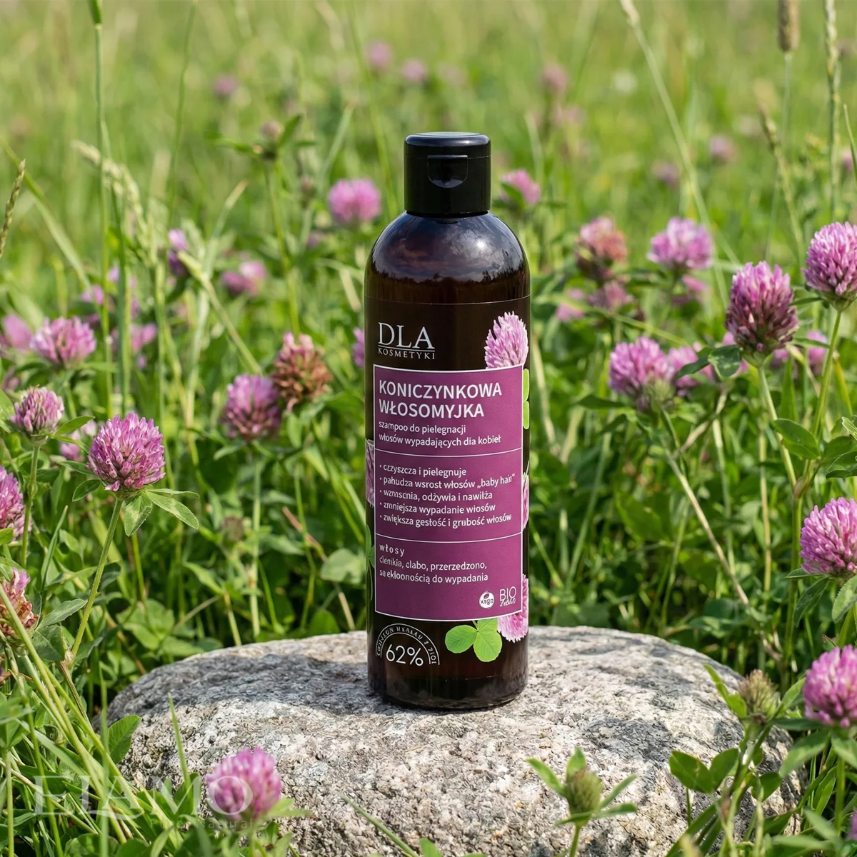 Butelka naturalnego szamponu "Koniczynkowa Włosomyjka" marki DLA Kosmetyki, stojąca na kamieniu na łące pełnej kwitnącej czerwonej koniczyny.