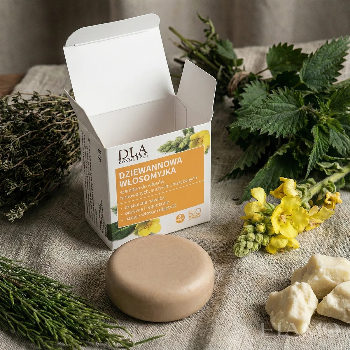 Rustykalna aranżacja na lnianym obrusie: otwarte opakowanie i kostka szamponu Dziewannowa Włosomyjka w otoczeniu świeżej pokrzywy, suszonego skrzypu, żółtych kwiatów dziewanny w wazonie oraz miseczki z masłem shea.