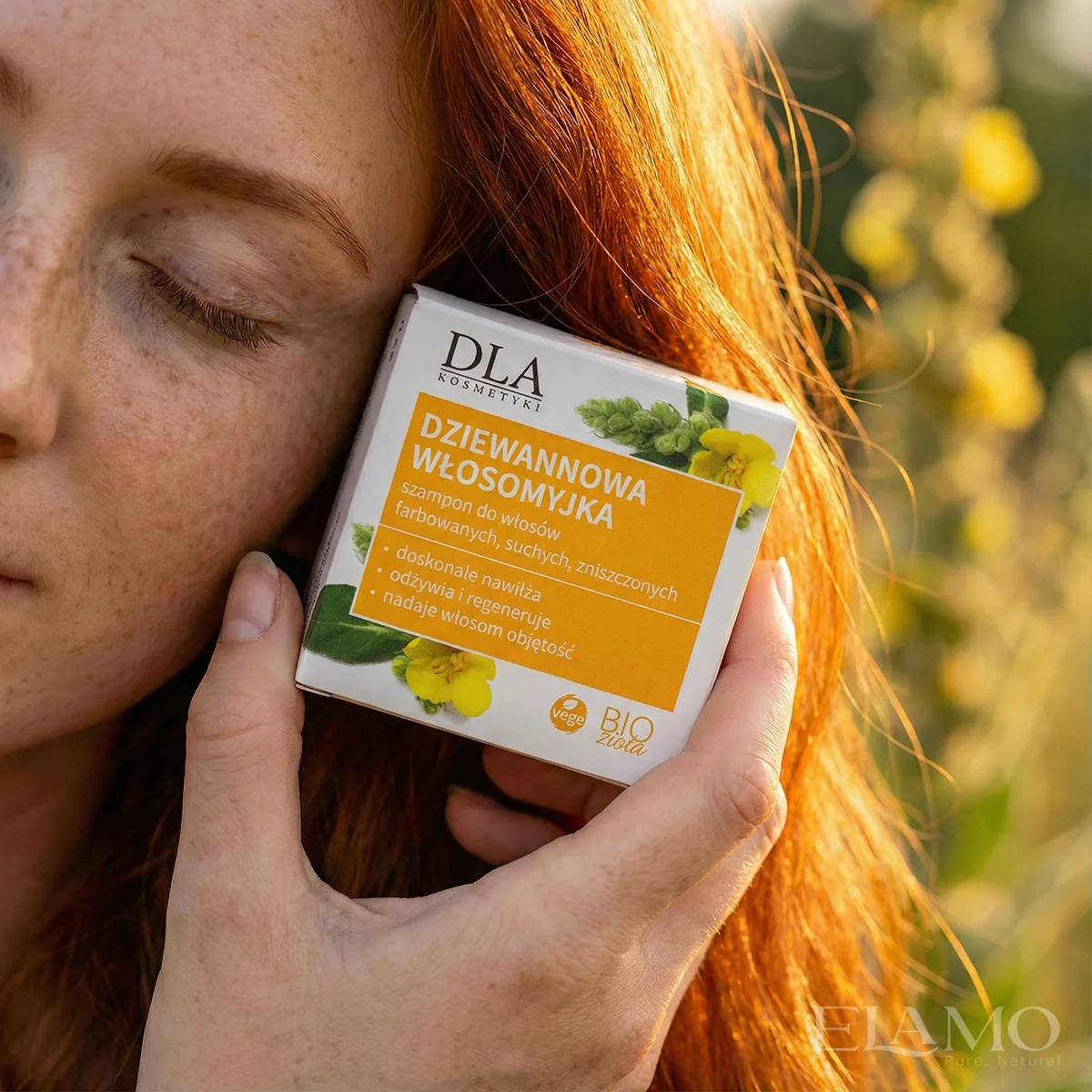 Rudowłosa modelka trzymająca przy policzku opakowanie szamponu Dziewannowa Włosomyjka w świetle zachodzącego słońca.