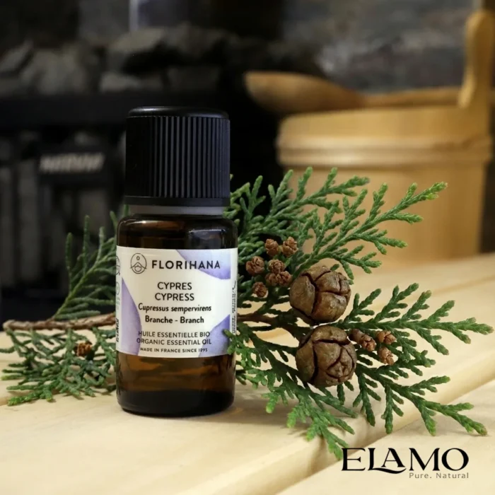 Organiczny olejek eteryczny Cyprys (Cypress) marki Florihana stojący na drewnianej ławie w saunie, obok naturalnej gałązki cyprysu z małymi szyszkami.