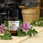 Duży olejek eteryczny Geranium Różane 15ml marki Florihana w scenerii sauny; w tle piec z kamieniami i drewniany ceber.
