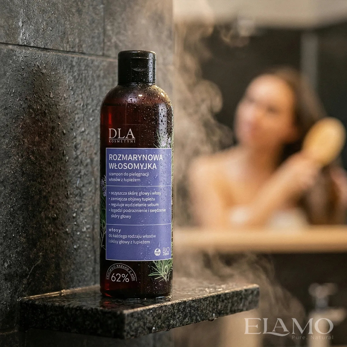 Butelka Rozmarynowej Włosomyjki pod prysznicem na mokrym kamieniu, w tle rozmyta postać kobiety czeszącej włosy.