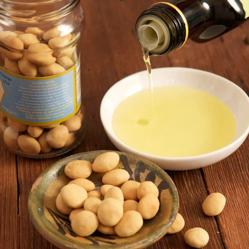 Organiczny olej z orzechów Sacha Inchi nalewany do białej miseczki obok ceramicznej misy z surowymi orzechami Sacha Inchi i zamkniętego słoika z etykietą informacyjną na drewnianym stole, bogate źródło kwasów Omega-3, 6, 9.