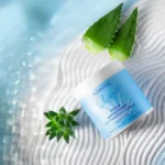 Zdjęcie opakowania kremu barierowego ALOESOVE BLUE do twarzy i ciała na tle falowanej, błękitnej tkaniny z naturalnymi liśćmi aloesu, podkreślającym intensywne nawilżenie i ochronę skóry.