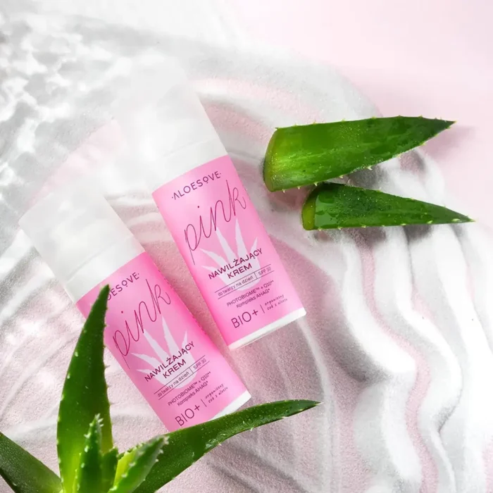 Zdjęcie przedstawiające dwa opakowania różowego nawilżającego kremu do twarzy ALOESOVE Pink z SPF 30, umieszczone na miękkiej, różowej tkaninie z liśćmi aloesu w tle.