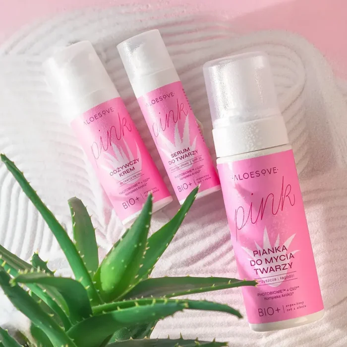 zdjęcie trzech różowych produktów kosmetycznych ALOESOVE Pink, w tym odżywczy krem do twarzy, serum do twarzy i pianka do mycia twarzy, ustawione na różowej, falistej tkaninie. W tle widoczne są soczyste, zielone liście aloesu.