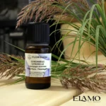 Organiczny olejek eteryczny Citronella Florihana na drewnianej ławie w saunie, udekorowany źdźbłami trawy cytrynowej ; w tle piec saunowy.