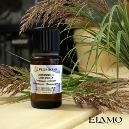 Organiczny olejek eteryczny Citronella Florihana na drewnianej ławie w saunie, udekorowany źdźbłami trawy cytrynowej ; w tle piec saunowy.