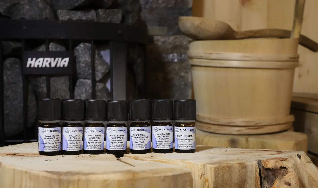 Naturalne olejki eteryczne do sauny – Florihana (Lavandula angustifolia, Eucalyptus globulus, Pinus sylvestris, Picea mariana, Abies balsamea, Cedrus atlantica, Ravintsara) na drewnie; w tle piec Harvia i cebrzyk.