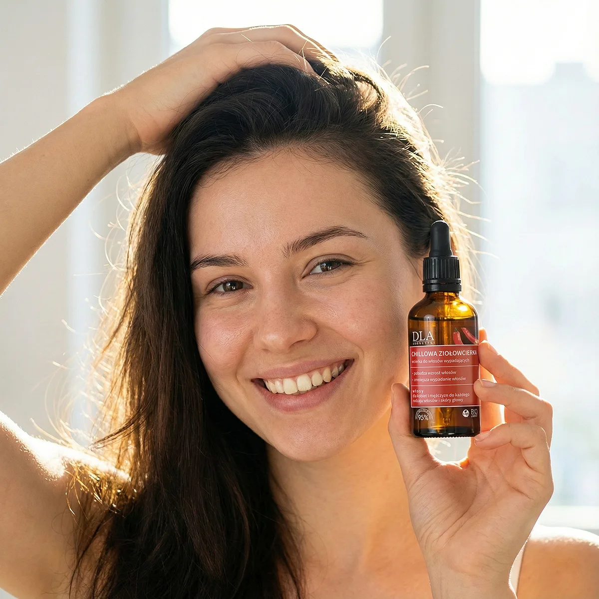 Uśmiechnięta kobieta z gęstymi, lśniącymi włosami trzymająca przy twarzy butelkę produktu Chillowa ziołowcierka.