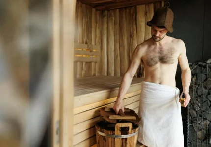 Sauna sucha – mężczyzna przygotowuje wodę polewową z rozcieńczonym naturalnym olejkiem eterycznym w drewnianym wiadrze; obok piec z kamieniami.