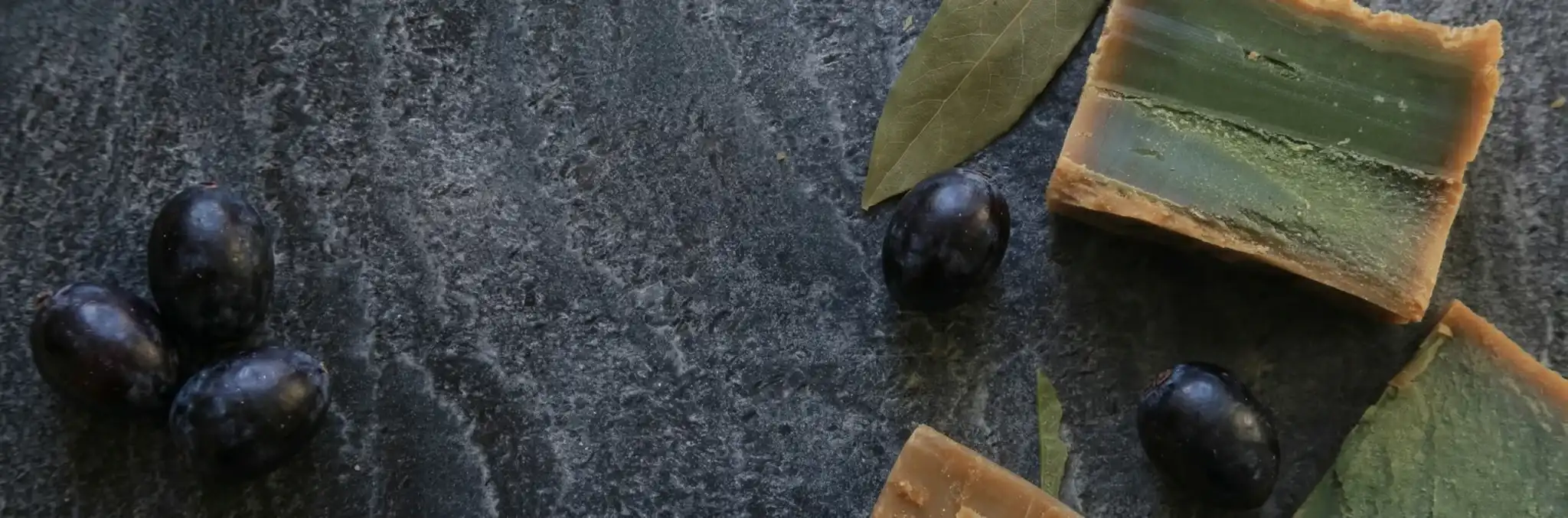 Oryginalne, ręcznie cięte kostki mydła Aleppo z widocznym zielonym wnętrzem, świeżymi oliwkami i liśćmi laurowymi na ciemnym tle